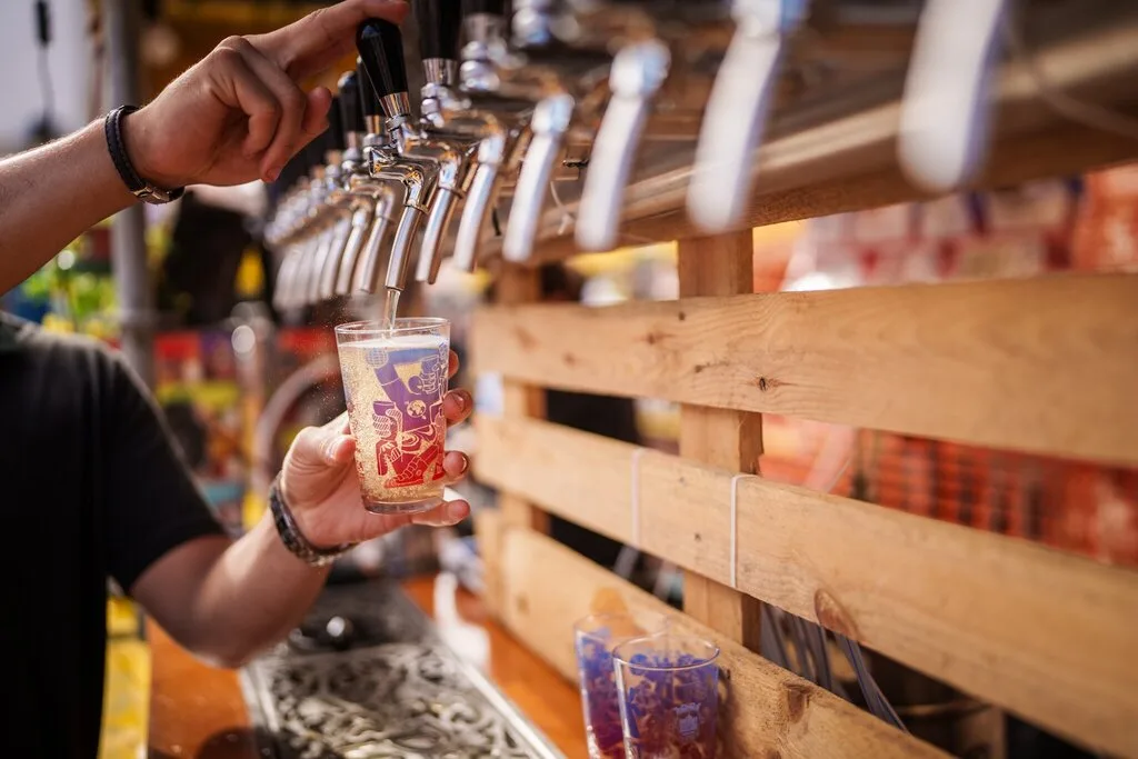 Mão enche um copo de cerveja artesanal numa torneira, com paletes de madeira ao fundo