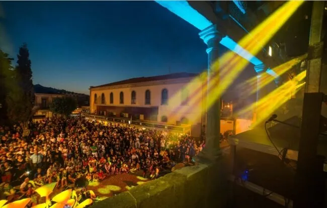 Multidão ao entardecer assiste a um concerto, com feixes de luz coloridos vindos do palco